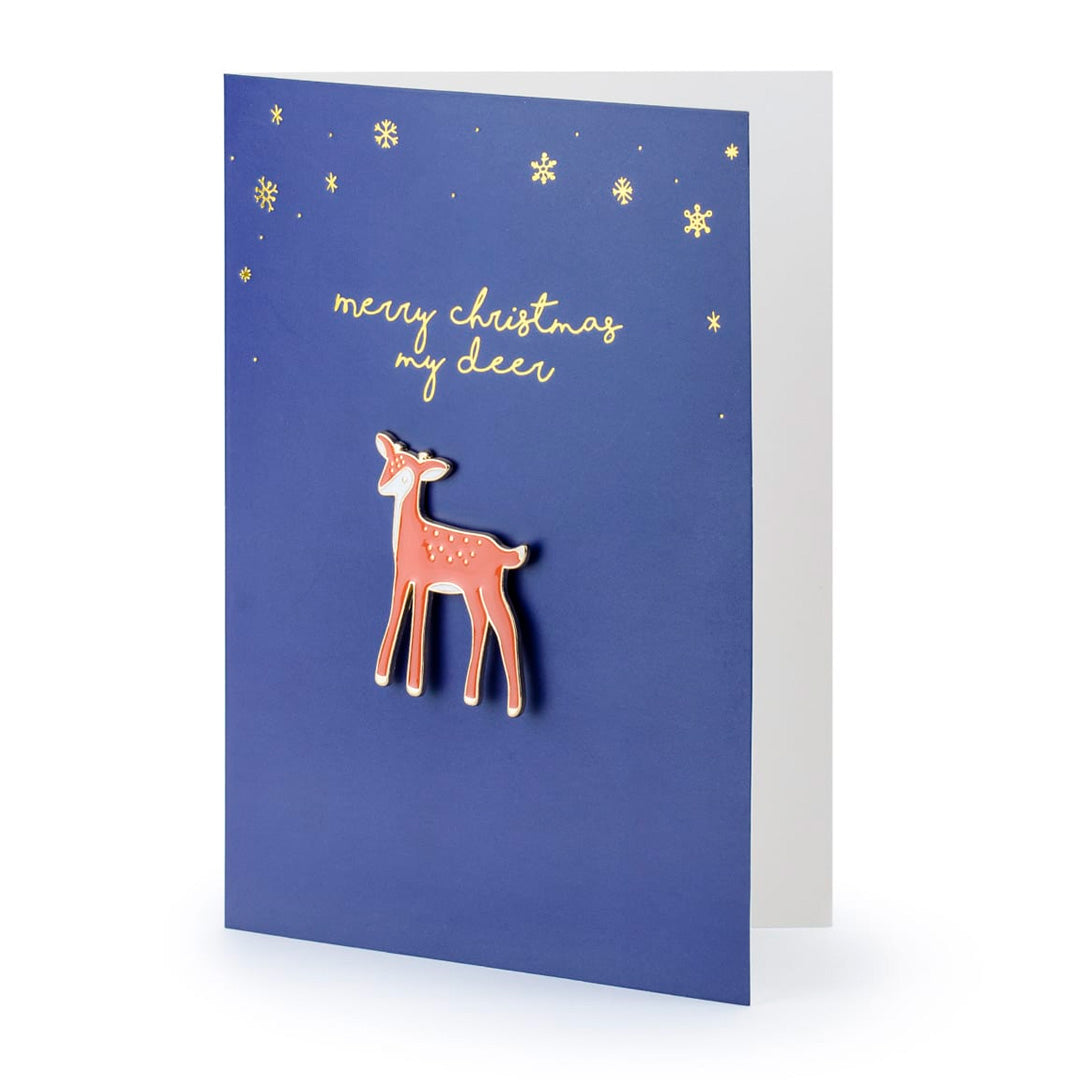 Detalle del pin de ciervo sobre tarjeta navideña azul con dorado
